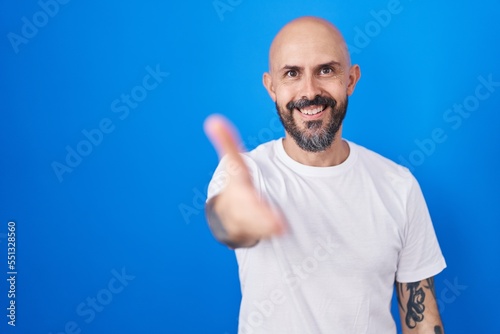 Hispanic man with tattoos standing over blue background smiling friendly offering handshake as greeting and welcoming. successful business.