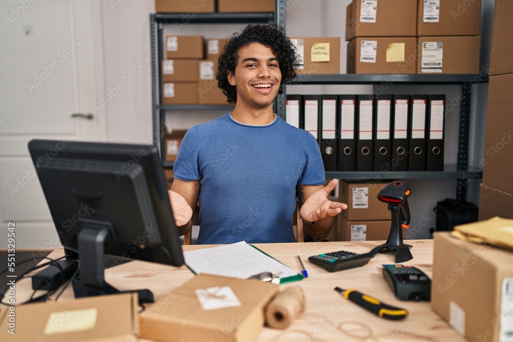 Hispanic man with curly hair working at small business ecommerce smiling cheerful with open arms as friendly welcome, positive and confident greetings