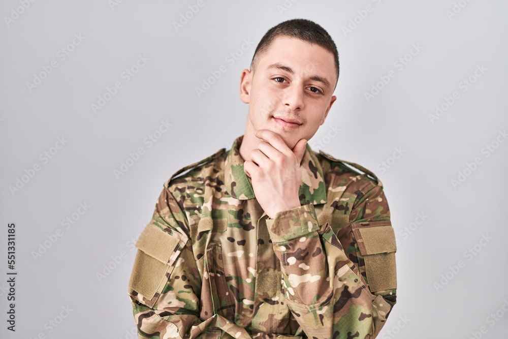 Young man wearing camouflage army uniform looking confident at the camera with smile with crossed arms and hand raised on chin. thinking positive. Young man wearing camouflage army uniform looking confident at the camera with smile with crossed arms and hand raised on chin. thinking positive.