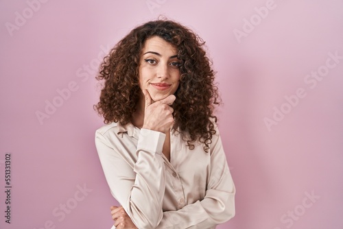 Hispanic woman with curly hair standing over pink background looking confident at the camera smiling with crossed arms and hand raised on chin. thinking positive.