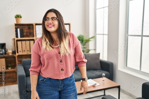 Young hispanic woman working at the office wearing glasses looking to side, relax profile pose with natural face and confident smile.