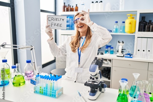 Young blonde scientist woman working on cruelty free laboratory smiling happy doing ok sign with hand on eye looking through fingers
