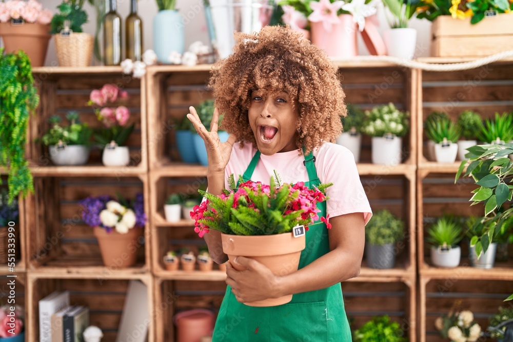 Young hispanic woman with curly hair working at florist shop holding plant celebrating victory with happy smile and winner expression with raised hands