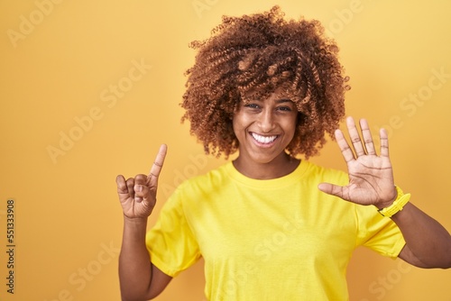 Young hispanic woman with curly hair standing over yellow background showing and pointing up with fingers number six while smiling confident and happy.
