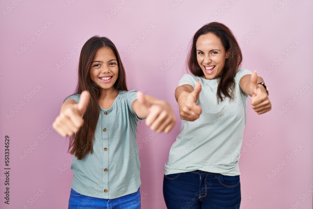 Young mother and daughter standing over pink background approving doing positive gesture with hand, thumbs up smiling and happy for success. winner gesture.