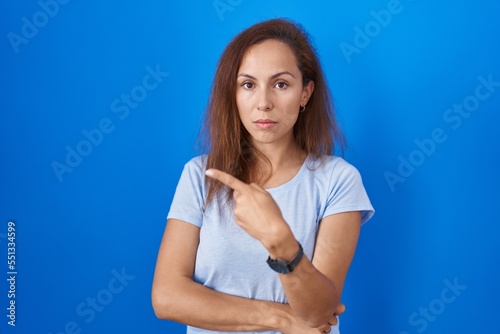 Brunette woman standing over blue background pointing with hand finger to the side showing advertisement, serious and calm face © Krakenimages.com
