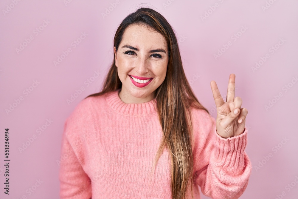 Young hispanic woman standing over pink background showing and pointing up with fingers number two while smiling confident and happy.