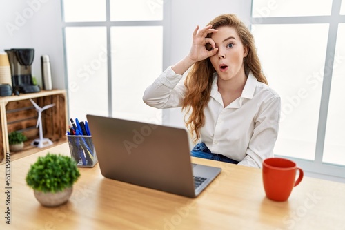 Young caucasian woman working at the office using computer laptop doing ok gesture shocked with surprised face, eye looking through fingers. unbelieving expression.