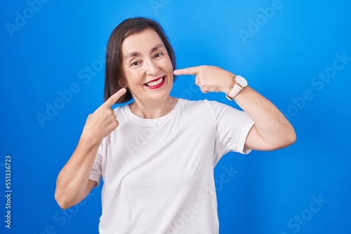 Middle age hispanic woman standing over blue background smiling cheerful showing and pointing with fingers teeth and mouth. dental health concept.