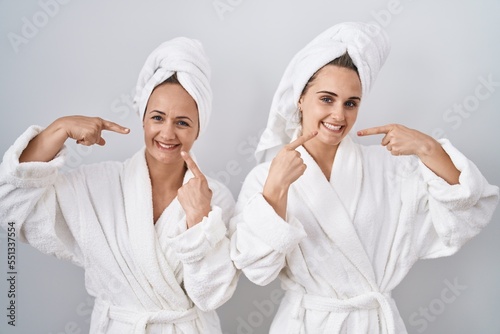 Middle age woman and daughter wearing white bathrobe and towel smiling cheerful showing and pointing with fingers teeth and mouth. dental health concept.
