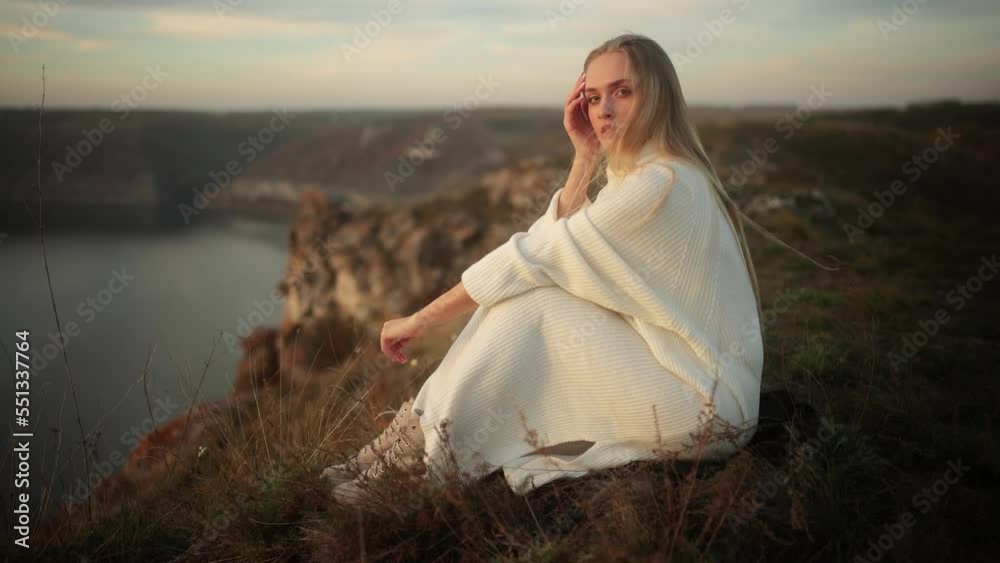 Beautiful young blonde woman in a long white knitted sweater sits on top of a mountain above a river and enjoys the sunset Beautiful young blonde woman in a long white knitted sweater sits on top of a mountain above a river and enjoys the sunset