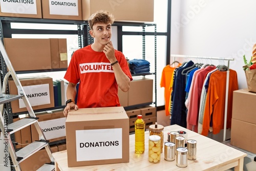 Young caucasian man volunteer holding donations box with hand on chin thinking about question, pensive expression. smiling and thoughtful face. doubt concept.