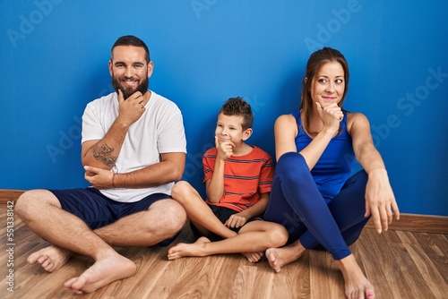 Family of three sitting on the floor at home looking confident at the camera smiling with crossed arms and hand raised on chin. thinking positive.