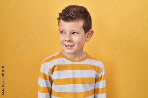 Young caucasian kid standing over yellow background looking away to side with smile on face, natural expression. laughing confident.