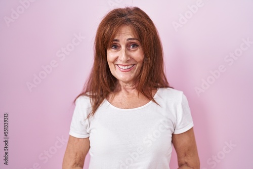 Middle age woman standing over pink background with hands together and crossed fingers smiling relaxed and cheerful. success and optimistic