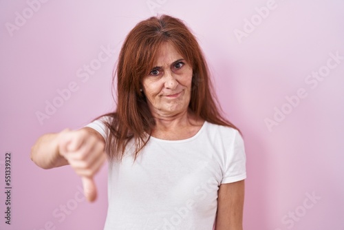Middle age woman standing over pink background looking unhappy and angry showing rejection and negative with thumbs down gesture. bad expression.