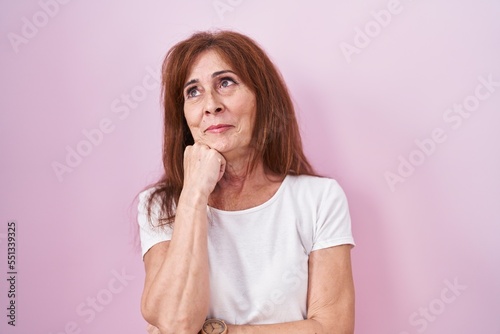 Middle age woman standing over pink background with hand on chin thinking about question, pensive expression. smiling with thoughtful face. doubt concept.