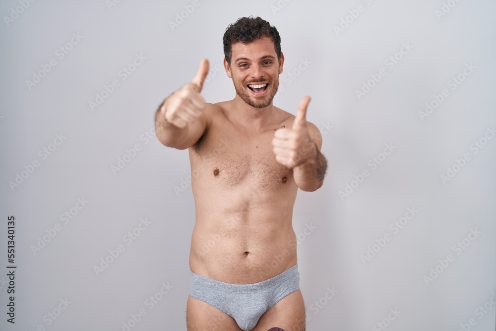 Fototapeta premium Young hispanic man standing shirtless wearing underware approving doing positive gesture with hand, thumbs up smiling and happy for success. winner gesture.