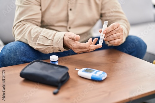 Middle age grey-haired man measuring glucose sitting on sofa at home