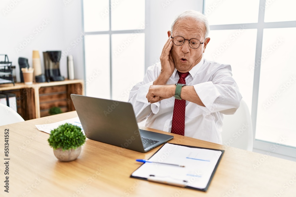 Senior man working at the office using computer laptop looking at the watch time worried, afraid of getting late Senior man working at the office using computer laptop looking at the watch time worried, afraid of getting late