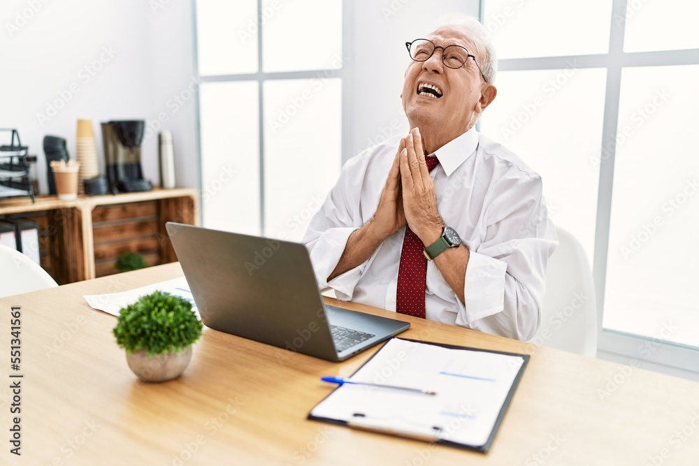 Senior man working at the office using computer laptop begging and praying with hands together with hope expression on face very emotional and worried. begging. Senior man working at the office using computer laptop begging and praying with hands together with hope expression on face very emotional and worried. begging.