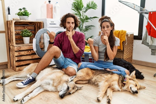 Photography Young hispanic couple doing laundry with dogs asking to be quiet with finger on lips pointing with hand to the side