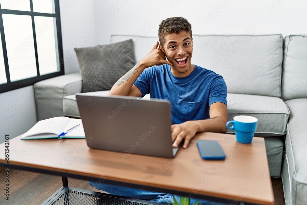 Young handsome hispanic man using laptop sitting on the floor smiling doing phone gesture with hand and fingers like talking on the telephone. communicating concepts. Young handsome hispanic man using laptop sitting on the floor smiling doing phone gesture with hand and fingers like talking on the telephone. communicating concepts.