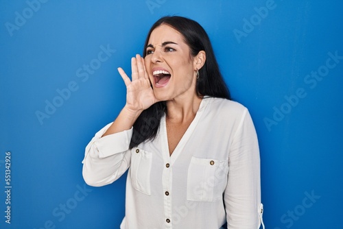 Young hispanic woman standing over blue background shouting and screaming loud to side with hand on mouth. communication concept.