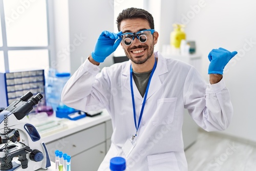 Young hispanic man working at scientist laboratory wearing magnifying glasses pointing thumb up to the side smiling happy with open mouth