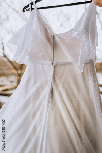 A beautiful long white bridesmaid dress hangs on a hanger in the morning outdoors. Photography, wedding, corset back.