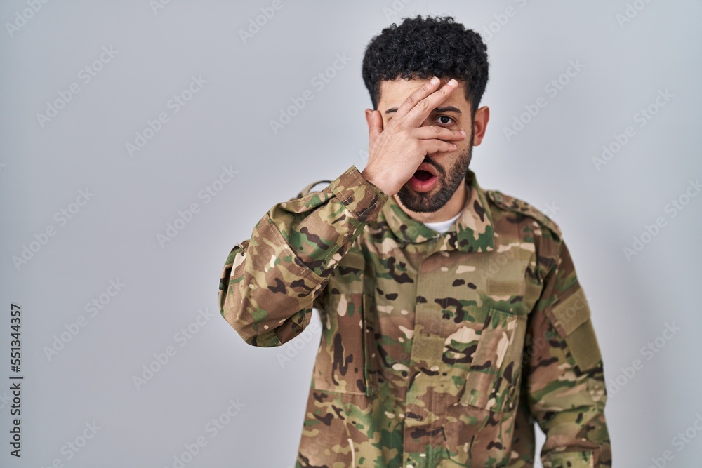 Arab man wearing camouflage army uniform peeking in shock covering face and eyes with hand, looking through fingers with embarrassed expression.