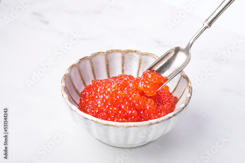 Red caviar in a beautiful ceramic bowl in the form of a flower with a silver spoon on a white  marble background. Seafood delicacies