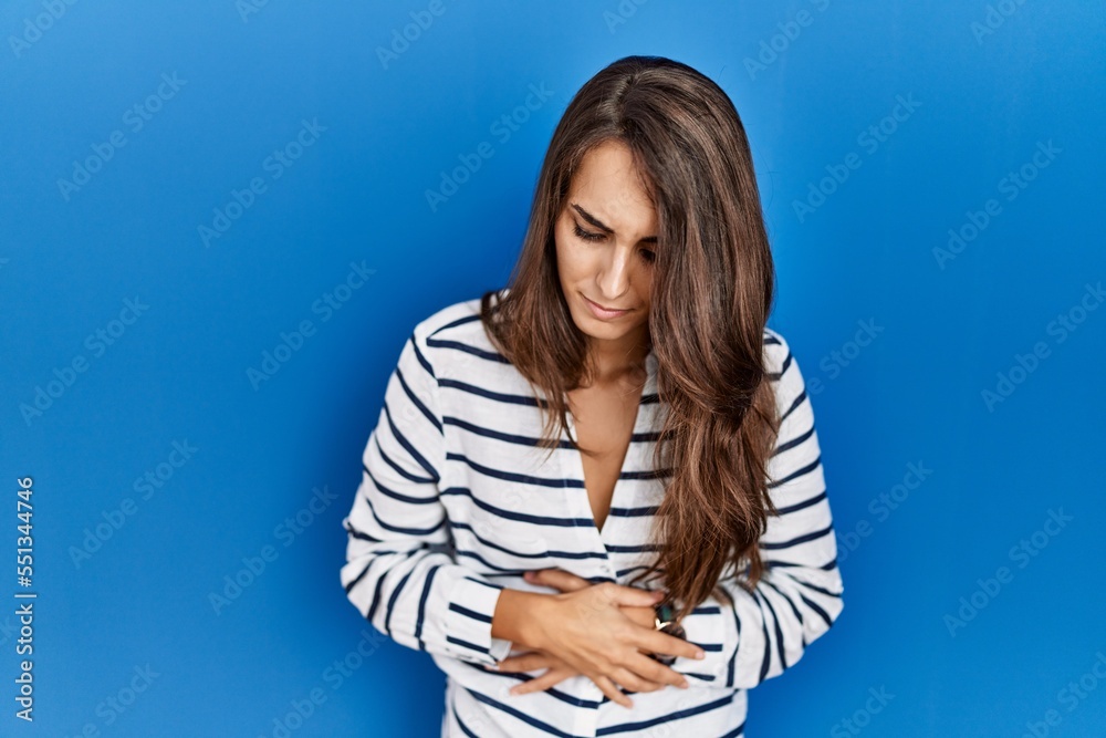 Young hispanic woman standing over blue isolated background with hand on stomach because indigestion, painful illness feeling unwell. ache concept. Young hispanic woman standing over blue isolated background with hand on stomach because indigestion, painful illness feeling unwell. ache concept.