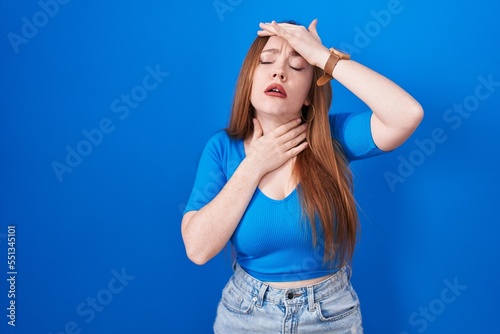 Redhead woman standing over blue background touching forehead for illness and fever, flu and cold, virus sick