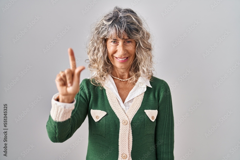 Middle age woman standing over white background showing and pointing up with finger number one while smiling confident and happy. Middle age woman standing over white background showing and pointing up with finger number one while smiling confident and happy.