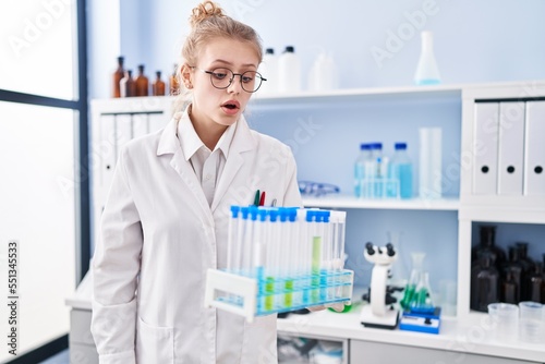 Young caucasian woman working at scientist laboratory holding samples scared and amazed with open mouth for surprise, disbelief face