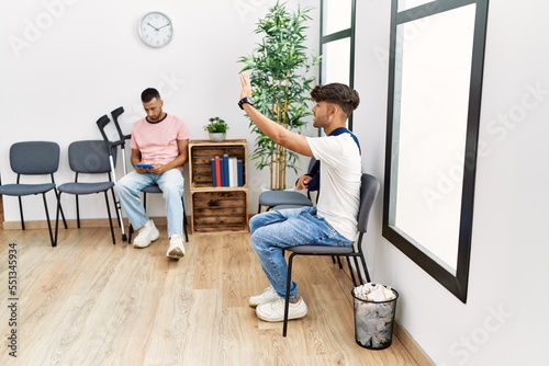 Two hispanic men using tablet sitting on chair at hospital waiting room photo