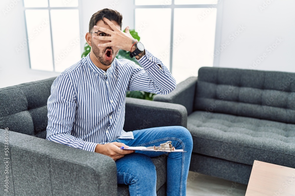 Young psychologist man at consultation office peeking in shock covering face and eyes with hand, looking through fingers with embarrassed expression. Young psychologist man at consultation office peeking in shock covering face and eyes with hand, looking through fingers with embarrassed expression.