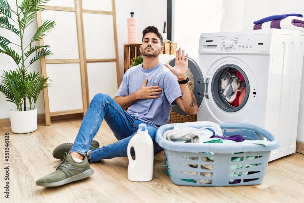 Young hispanic man putting dirty laundry into washing machine swearing with hand on chest and open palm, making a loyalty promise oath Young hispanic man putting dirty laundry into washing machine swearing with hand on chest and open palm, making a loyalty promise oath