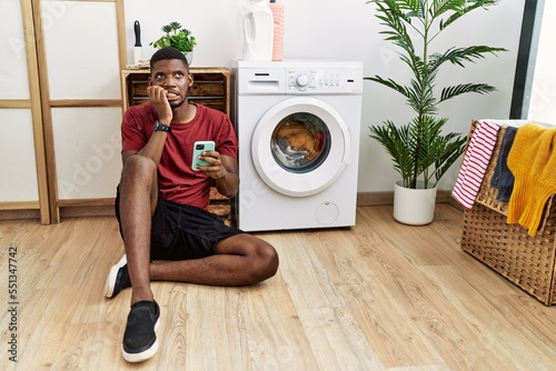 Young african american man using smartphone waiting for washing machine looking stressed and nervous with hands on mouth biting nails. anxiety problem. photo