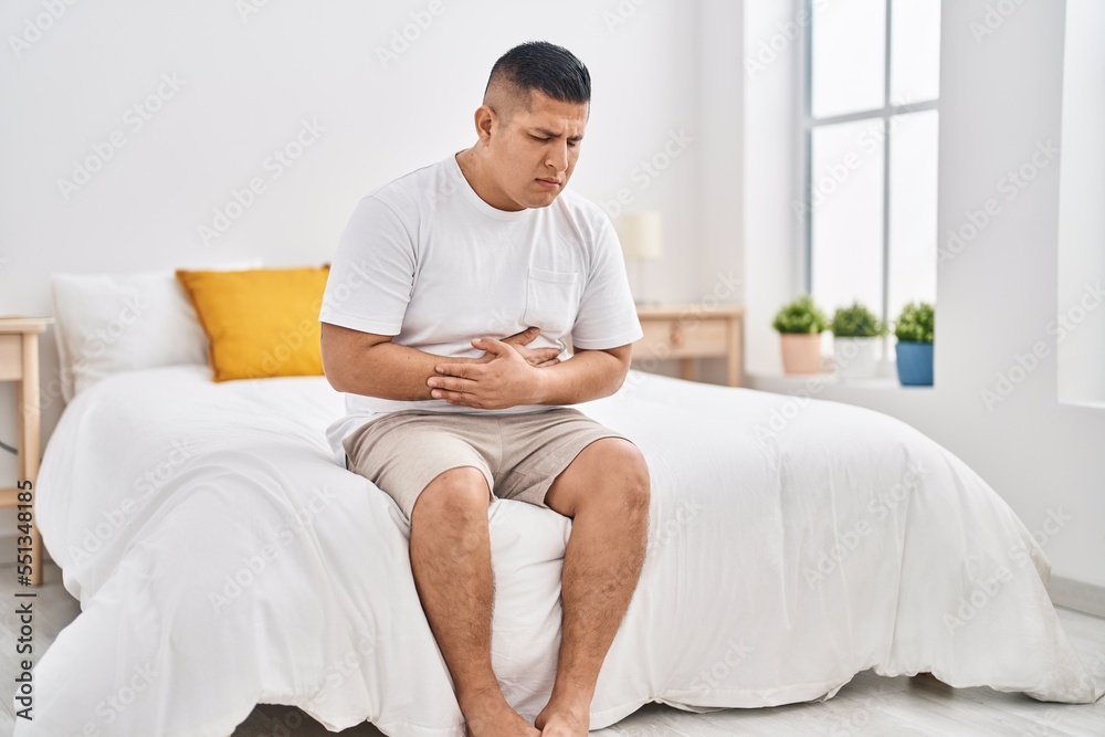 Young latin man suffering for stomach ache sitting on bed at bedroom Young latin man suffering for stomach ache sitting on bed at bedroom