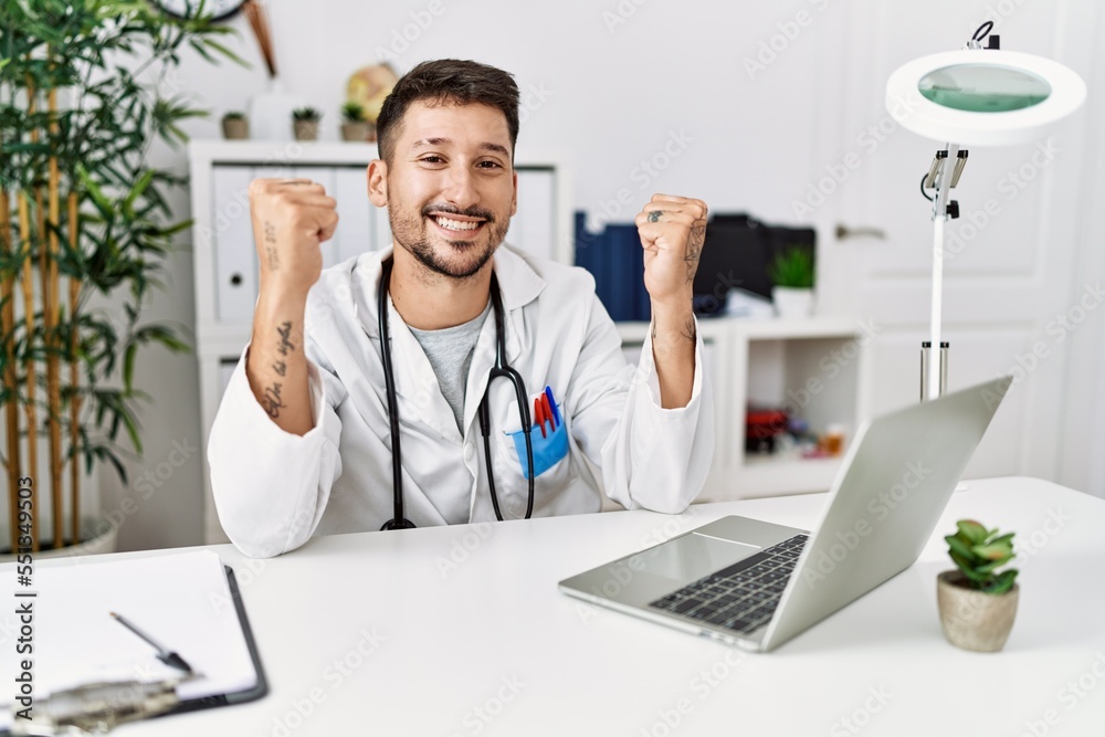 Young doctor working at the clinic using computer laptop very happy and excited doing winner gesture with arms raised, smiling and screaming for success. celebration concept.