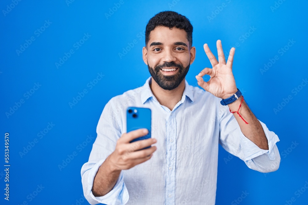 Hispanic man with beard using smartphone typing message smiling positive doing ok sign with hand and fingers. successful expression. Hispanic man with beard using smartphone typing message smiling positive doing ok sign with hand and fingers. successful expression.