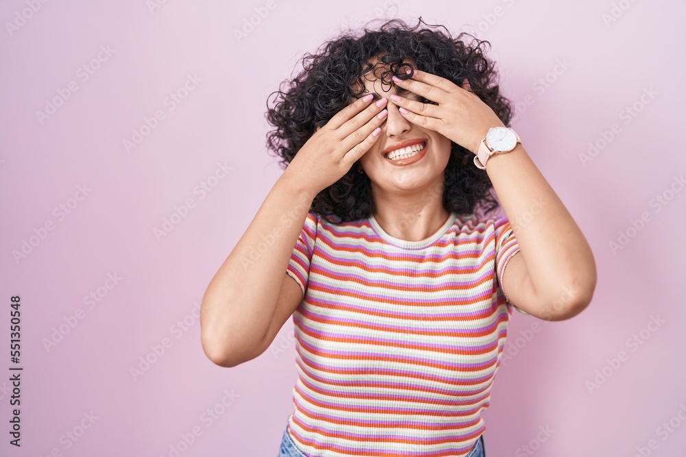 Young middle east woman standing over pink background covering eyes with hands smiling cheerful and funny. blind concept.