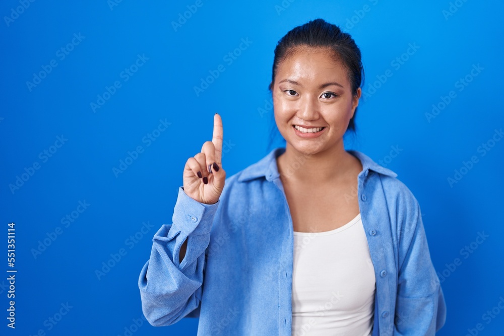 Asian young woman standing over blue background showing and pointing up with finger number one while smiling confident and happy. Asian young woman standing over blue background showing and pointing up with finger number one while smiling confident and happy.