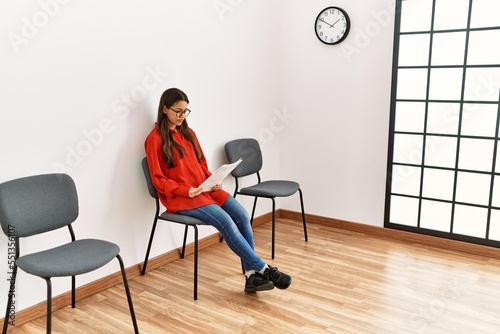 Young latin woman reading document sitting on chair at waiting room