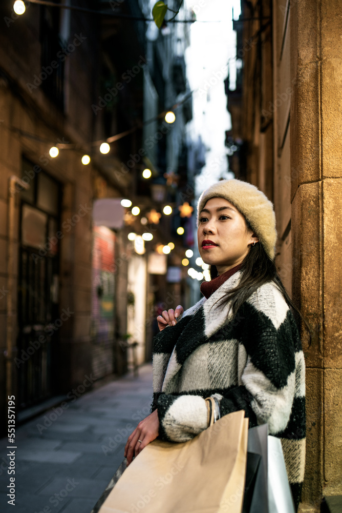 Pensive Chinese female in outerwear and beret with shopping bags in hands standing on street near residential buildings in city