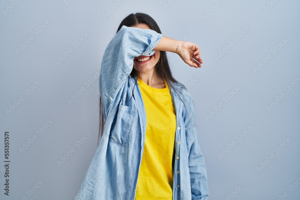 Young hispanic woman standing over blue background covering eyes with arm smiling cheerful and funny. blind concept. Young hispanic woman standing over blue background covering eyes with arm smiling cheerful and funny. blind concept.
