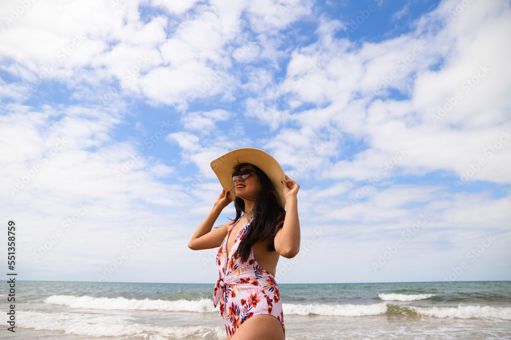 Beautiful young woman posing on the beach with a hat to protect herself from the sun. The woman is enjoying her trip to a paradise beach. Holiday and travel concept.