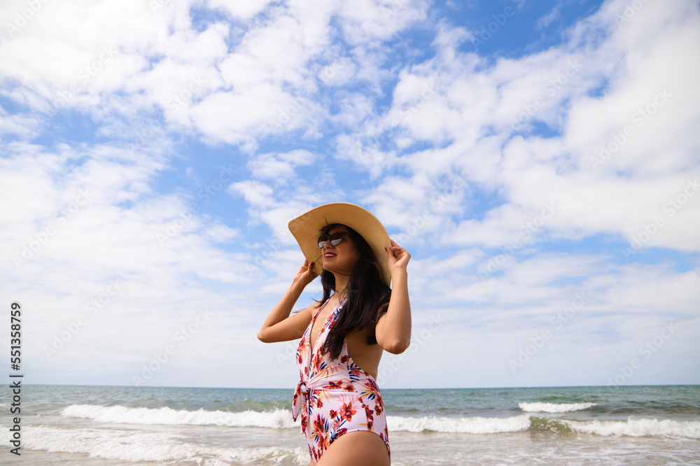 Beautiful young woman posing on the beach with a hat to protect herself from the sun. The woman is enjoying her trip to a paradise beach. Holiday and travel concept. Beautiful young woman posing on the beach with a hat to protect herself from the sun. The woman is enjoying her trip to a paradise beach. Holiday and travel concept.
